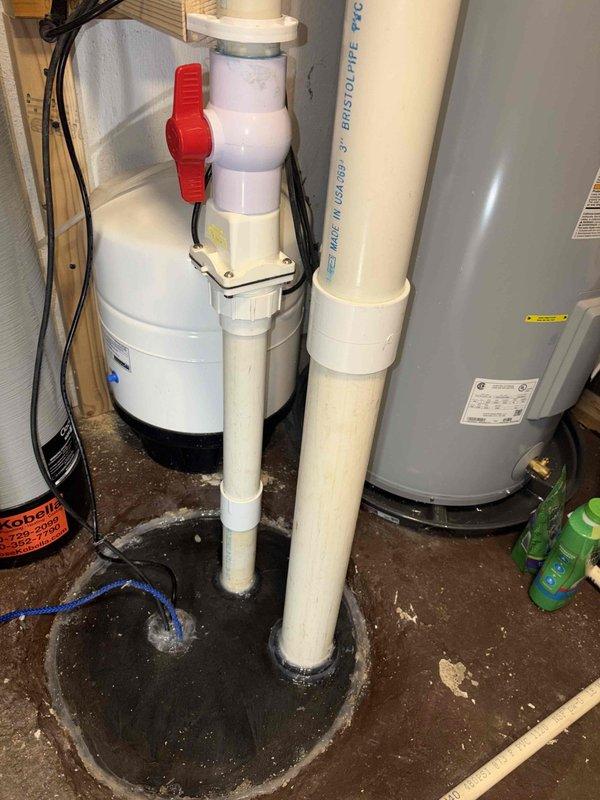 Completed sump pump installation in basement mechanical room. Installed white cylindrical sump basin with dual PVC discharge piping system, including SDR 35 PVC DWV pipe with ball valve for system control. Routed discharge lines vertically through floor penetration with proper weatherproof sealing boots and waterproofing compound applied at penetration points. System is operational with water visible in basin indicating proper drainage function. Customer advised that return visit is scheduled to install basin lid to eliminate odor issues from open sump pit.