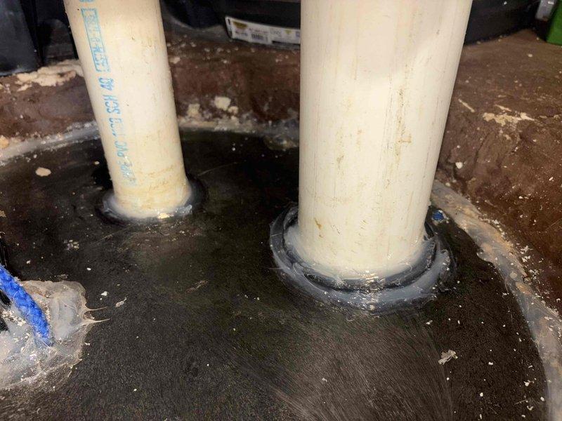 Completed sump pump installation in basement mechanical room. Installed white cylindrical sump basin with dual PVC discharge piping system, including SDR 35 PVC DWV pipe with ball valve for system control. Routed discharge lines vertically through floor penetration with proper weatherproof sealing boots and waterproofing compound applied at penetration points. System is operational with water visible in basin indicating proper drainage function. Customer advised that return visit is scheduled to install basin lid to eliminate odor issues from open sump pit.