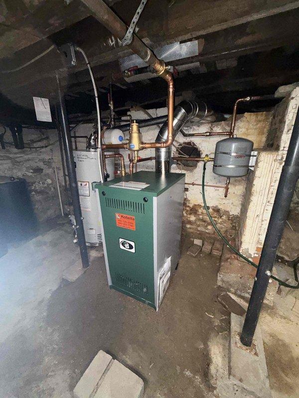 Completed boiler replacement in basement mechanical room, removing existing green boiler unit and associated equipment with assistance of additional technician and power dolly as required. Installed new boiler system with proper connections to existing copper piping, brass fittings, and distribution lines extending to overhead joists. Verified all connections to water heater, expansion tank, and related plumbing components for proper integration with new heating system.