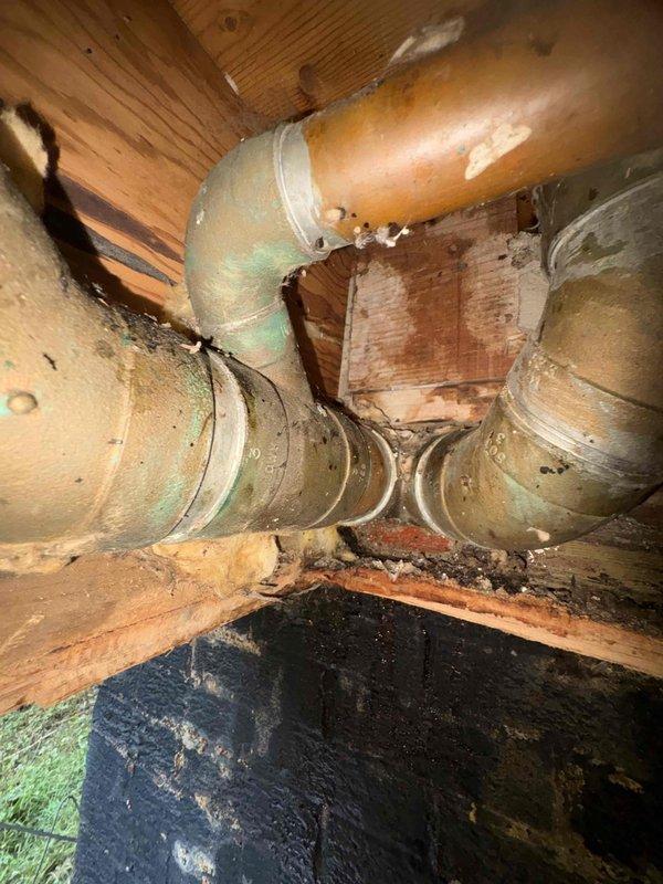 Responded to water leak in basement bathroom area. Upon inspection, identified active leak at PVC to copper pipe transition point. The copper piping system showed significant age-related deterioration with green patina oxidation and corrosion present throughout multiple sections. Leak was isolated to the coupling joint where white PVC pipe connects to copper supply line. Repaired transition connection using appropriate coupling hardware and secured with metal clamp. Verified repair integrity and confirmed no additional leaks present in surrounding plumbing system.
