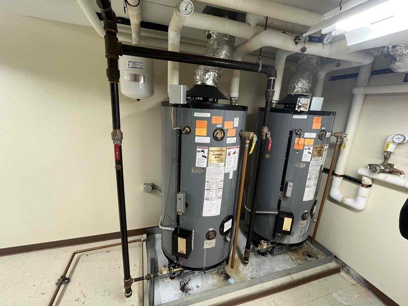 Completed on-site assessment for replacement of two 100-gallon commercial water heaters currently installed on raised platform in mechanical room. Documented existing system configuration including dual tank setup with associated piping infrastructure, expansion tank, pressure gauges, and connection points to building's hot water distribution system. Photographed current installation showing PVC condensate drainage, copper supply lines, flexible gas connections, and HVAC integration components to provide comprehensive estimate for replacement equipment and labor requirements.