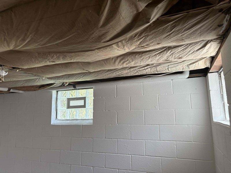 Completed site inspection of basement utility area to assess HVAC infrastructure and conditions. Documented exposed ceiling joists with fiberglass insulation installation, noting areas where batting material has sagged and requires repositioning for optimal thermal efficiency. Photographed PVC drainage lines running through wooden framing along concrete block walls to verify proper routing and clearances for future HVAC equipment considerations.