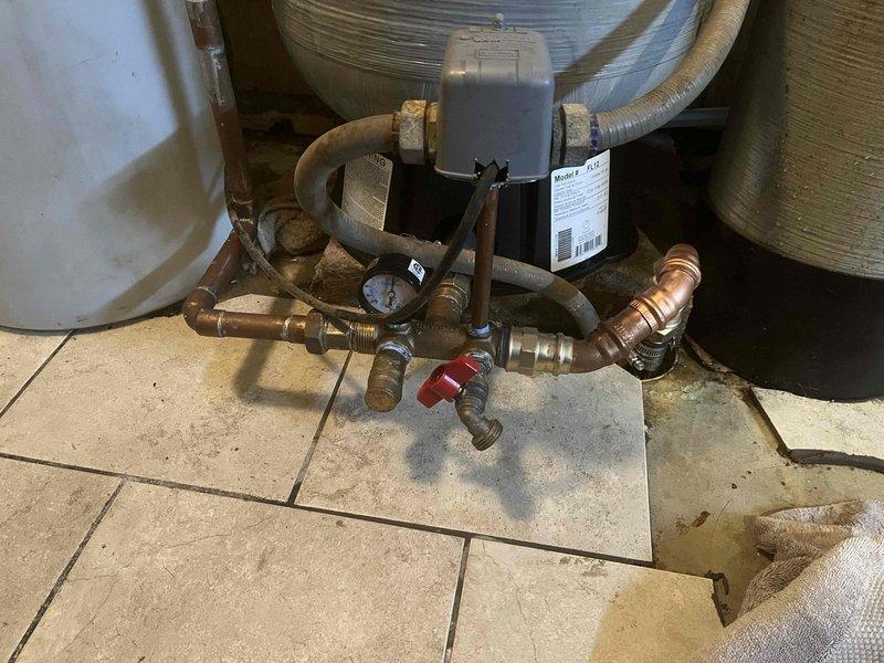 Responded to emergency call for major water leak inside fifty-year-old residence. Upon arrival, identified active leak at water heater connection point with water pooling on concrete floor beneath compromised pipe fitting. Applied temporary blue sealing tape to leaking connection to mitigate immediate water discharge. Inspected complete water heater plumbing assembly including brass manifold connections, pressure relief valve, shutoff valves, and insulated supply lines. Assessed condition of aged brass fittings showing oxidation and verified pressure gauge functionality. Documented existing installation configuration and leak source for repair recommendations. Controlled water flow to prevent further interior damage while evaluating system integrity.