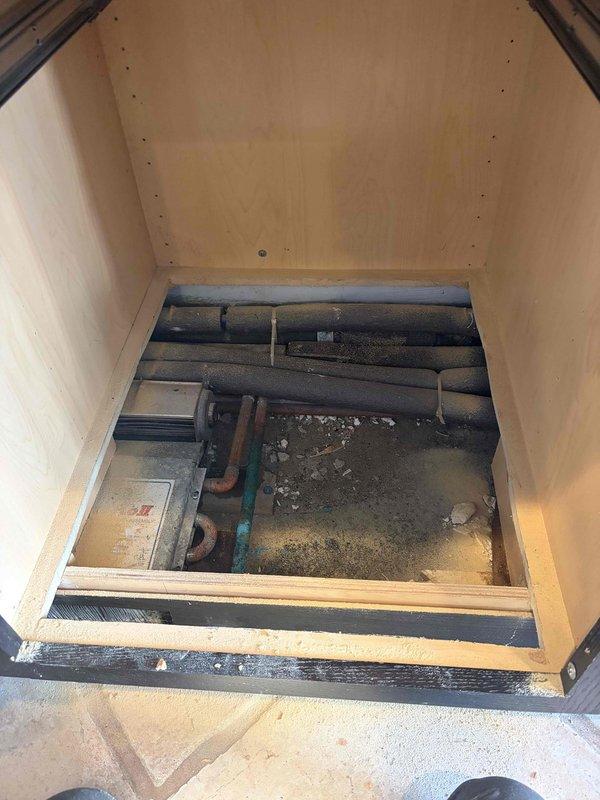 Responded to service call for leaking kitchen baseboard heater. Upon inspection, discovered active water leak from circulator pump assembly in wooden access compartment beneath kitchen floor. Found significant water accumulation in metal drain pan with evidence of ongoing moisture exposure and mineral deposits on surrounding surfaces. Documented conditions with photographs showing teal-colored pump component, associated piping with black insulation, and water pooling at base of access shaft.