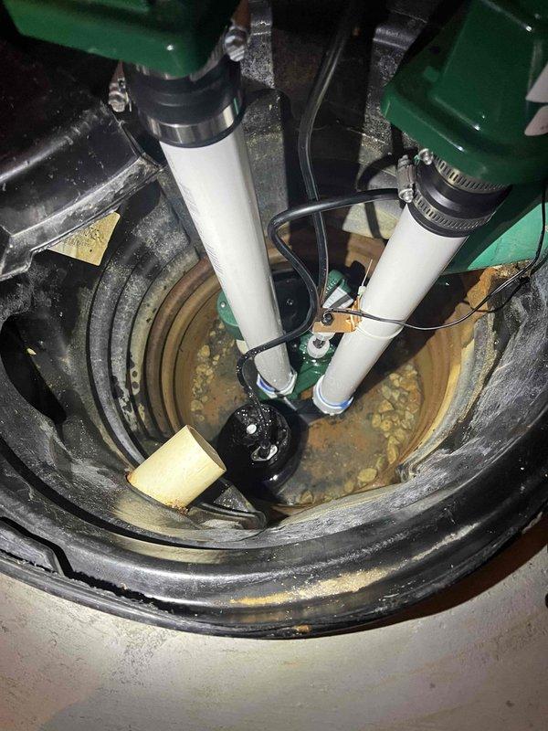 Completed comprehensive inspection of dual-pump sump system. Evaluated submersible pump operation, discharge piping configuration with white PVC and black flexible hoses, and overall basin condition. Documented significant sediment accumulation and debris in standing water at basin floor, both green-colored pumps operational and properly secured with metal clamps and strapping, and all connections intact. Photographed system for service records showing typical wear patterns consistent with regular use.