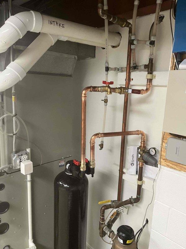 # Whole Home Descaler Installation

Completed return service visit for previously sold whole home descaler system. Installed comprehensive water treatment system including large-capacity black descaler tank with complete copper piping infrastructure and brass valve assembly. Configured system integration with existing A.O. Smith water heater unit, incorporating pressure gauges, red-handled isolation valves, and multi-point connection system. Established proper intake routing using white PVC piping with labeled components for easy identification. Installed auxiliary equipment including condensate management system with electrical connections. All copper pipe runs secured to wall mounting with professional-grade fittings and elbow joints throughout vertical and horizontal distribution network. Verified proper pressure readings, tested all valve operations, and confirmed system integration with existing plumbing infrastructure. Installation completed to manufacturer specifications with al