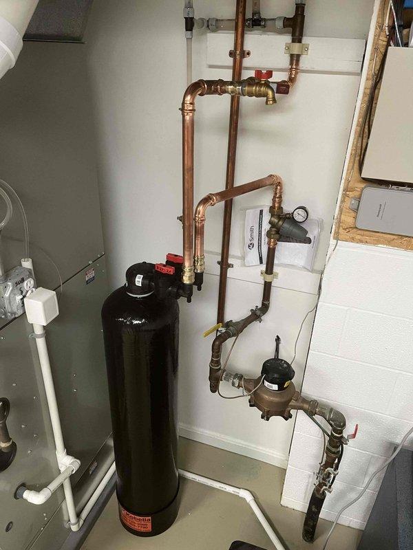# Whole Home Descaler Installation

Completed return service visit for previously sold whole home descaler system. Installed comprehensive water treatment system including large-capacity black descaler tank with complete copper piping infrastructure and brass valve assembly. Configured system integration with existing A.O. Smith water heater unit, incorporating pressure gauges, red-handled isolation valves, and multi-point connection system. Established proper intake routing using white PVC piping with labeled components for easy identification. Installed auxiliary equipment including condensate management system with electrical connections. All copper pipe runs secured to wall mounting with professional-grade fittings and elbow joints throughout vertical and horizontal distribution network. Verified proper pressure readings, tested all valve operations, and confirmed system integration with existing plumbing infrastructure. Installation completed to manufacturer specifications with al