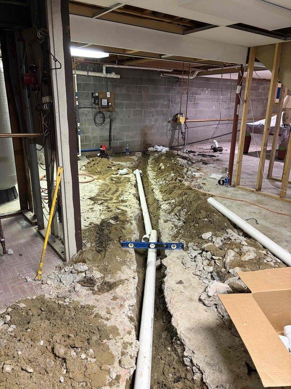 Returned to property to continue exterior clean-out installation previously sold. Excavated basement floor and installed parallel runs of white PVC drainage piping in prepared trenches, ensuring proper slope and alignment using leveling tools. Work is currently on hold pending completion of restoration activities by fire and water damage remediation company before final backfill and concrete restoration can be completed.