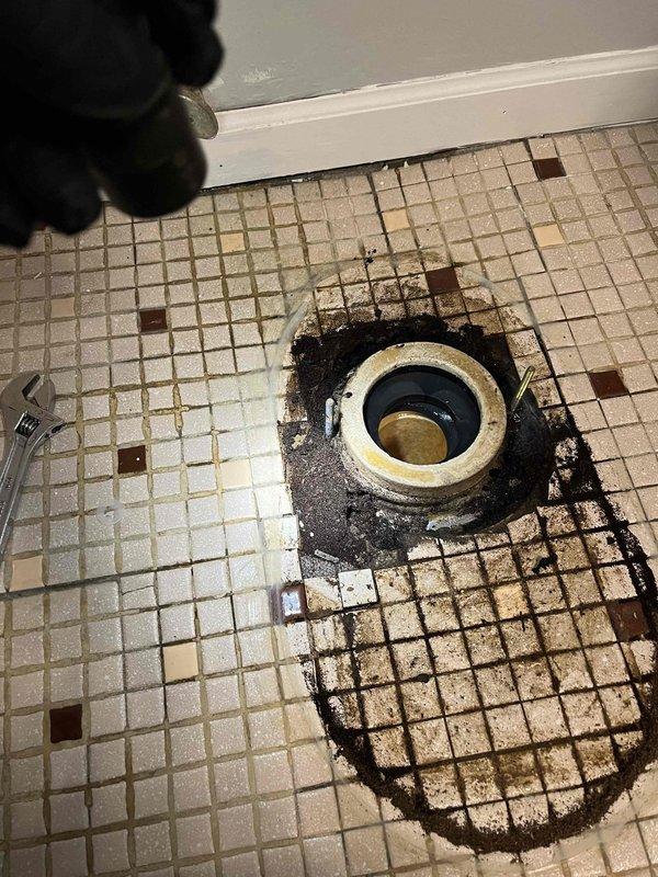 Responded to basement ceiling pipe leak near toilet. Upon inspection, discovered significant water damage to bathroom floor surrounding toilet flange, with extensive dark staining across mosaic tile flooring indicating prolonged moisture intrusion. Removed toilet to assess damage extent. Found deteriorated subflooring with broken and missing tiles around flange area, confirming active leak from overhead plumbing had compromised floor integrity. Documented existing yellow plastic toilet flange with rubber gasket and surrounding water-damaged tiles showing rot and structural deterioration requiring remediation before toilet reinstallation.