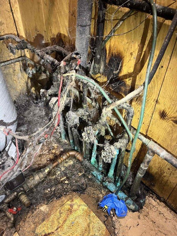 Responded to no water complaint at residential property. Upon inspection, discovered water treatment system with severe fire and heat damage affecting primary supply lines. Multiple copper supply pipes showed extensive corrosion, melting, and oxidation with characteristic green patina from extreme heat exposure. Electrical wiring to system controls exhibited burn damage with exposed sections. Water softener tank and associated components compromised by fire damage in utility area. Documented existing conditions showing charred wall surfaces, damaged piping infrastructure, and debris accumulation. System rendered inoperable due to extent of fire damage requiring complete replacement of affected plumbing components and water treatment equipment to restore water service.