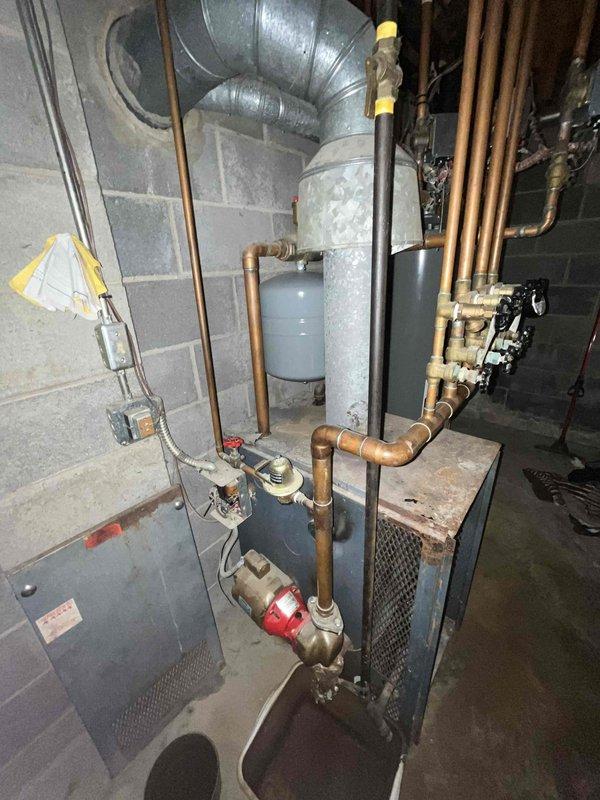 Responded to service call for water leak on aging furnace system in basement. Upon arrival, inspected 20+ year old heating unit and associated plumbing connections. Identified source of leak at copper piping connections to boiler unit. Examined red circulation pump, expansion tank, and water heater connections for additional moisture or compromised fittings. Documented existing conditions of mechanical systems including oxidized copper piping, brass valves, and plumbing manifold. Placed collection bucket beneath active leak point to prevent floor damage. Assessed overall condition of heating system and associated components. Due to age of equipment and extent of deterioration observed in piping and connections, recommended evaluation for system replacement versus repair options. Provided customer with photographic documentation of equipment condition and leak location for review and decision on next steps.