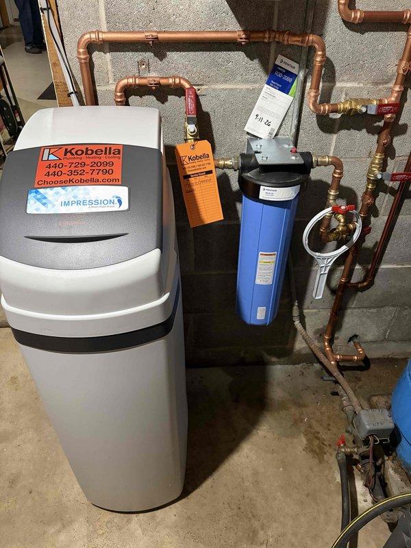 Completed installation of residential water treatment system including Impression water softener unit and blue cylindrical water filter housing with pressure gauge. Configured extensive copper piping network with multiple shut-off valves featuring red handles for maintenance access and system isolation, connecting treatment equipment to main water supply. All components mounted securely to concrete block wall with proper valve placement and professional plumbing connections utilizing brass fittings and compression joints throughout the system.