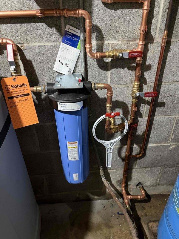 Completed installation of residential water treatment system including Impression water softener unit and blue cylindrical water filter housing with pressure gauge. Configured extensive copper piping network with multiple shut-off valves featuring red handles for maintenance access and system isolation, connecting treatment equipment to main water supply. All components mounted securely to concrete block wall with proper valve placement and professional plumbing connections utilizing brass fittings and compression joints throughout the system.
