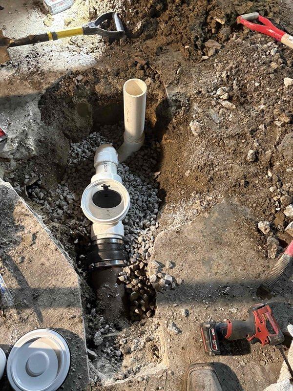 Addressed recurring floor drain backup issues by excavating and upgrading the drainage system. Removed existing floor drain 90-degree elbow configuration and installed new trap assembly with white PVC piping and proper drainage bedding. Excavation work included digging to expose existing plumbing infrastructure, installing vertical drain pipe with secure coupling connections, and backfilling with gravel drainage layer to ensure proper water flow and prevent future backup occurrences.