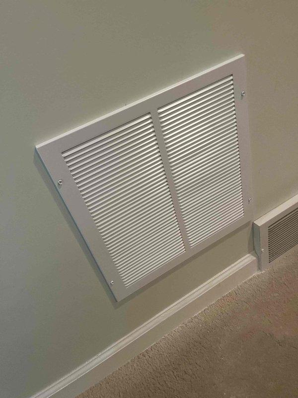 Installed return air ductwork connection for humidifier system in basement utility area. Connected flexible ducting from floor-mounted HVAC unit to rigid metal ductwork running along ceiling joists, with reflective metallic insulation wrap applied for thermal efficiency. Installed new white metal return air vent grille with horizontal slats on upper wall near ceiling to complete the air circulation pathway.