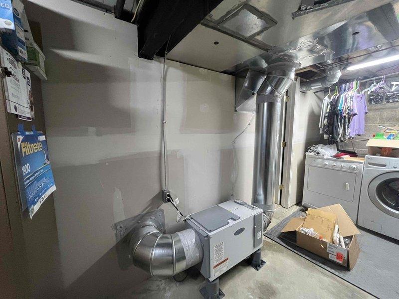 Installed return air ductwork connection for humidifier system in basement utility area. Connected flexible ducting from floor-mounted HVAC unit to rigid metal ductwork running along ceiling joists, with reflective metallic insulation wrap applied for thermal efficiency. Installed new white metal return air vent grille with horizontal slats on upper wall near ceiling to complete the air circulation pathway.