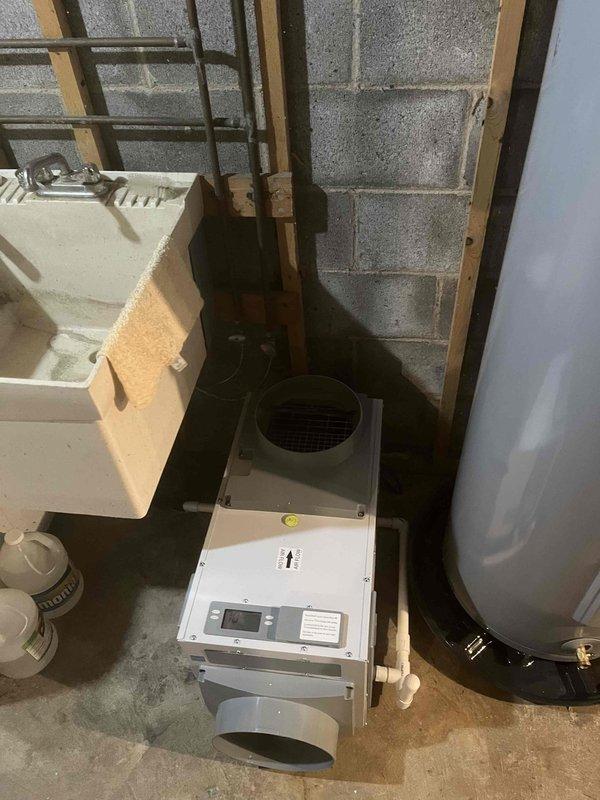Returned to property to complete bathroom drain repipe project and installed dehumidifier unit in basement utility area. Mounted and configured new dehumidifier system with digital control panel featuring ON/OFF and MODE controls, positioned unit on concrete floor adjacent to existing plumbing infrastructure. Connected ventilation ductwork to unit and integrated with existing HVAC system using flexible insulated ducting. Verified proper operation of control interface and display functionality upon installation completion.