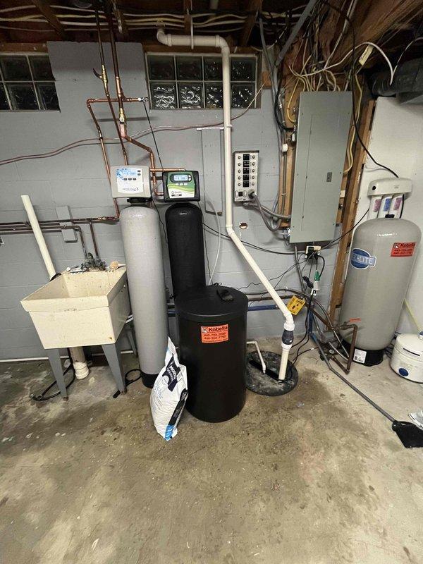 Completed comprehensive water treatment system installation including water softener, filtration equipment, and water heater in basement utility area. Installed tee connection for furnace humidifier with quarter-inch copper line run to hot water room with extra line left for future needs. Connected vinyl discharge tube from humidifier base to drain line to ensure proper condensate removal and system operation.