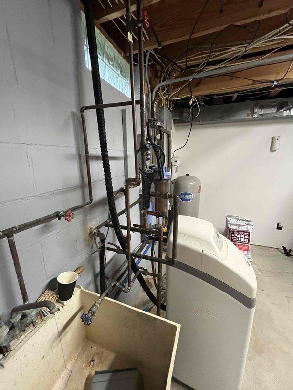 Completed comprehensive water treatment system installation including water softener, filtration equipment, and water heater in basement utility area. Installed tee connection for furnace humidifier with quarter-inch copper line run to hot water room with extra line left for future needs. Connected vinyl discharge tube from humidifier base to drain line to ensure proper condensate removal and system operation.