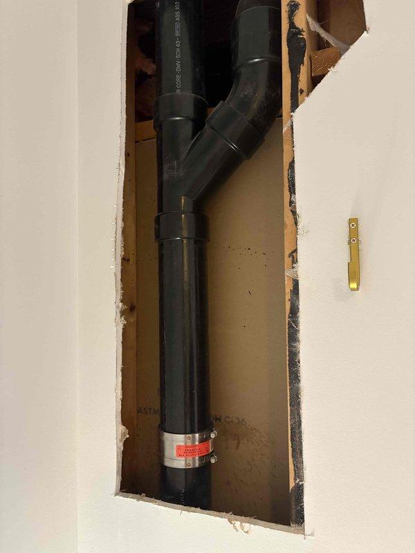 Completed return visit for pipe repair service previously sold. Accessed interior wall cavity through existing service panel to inspect and repair black PVC drainage system with Y-configuration. Removed and replaced defective plumbing components including threaded metallic cap, white plastic washer, and green plastic adapter fitting. Serviced T-shaped metal valve assembly with multiple connection points in utility area, ensuring proper installation of compression fittings and secure connections to flexible corrugated supply line. All replaced components tested for proper function and verified no leaks present upon completion.