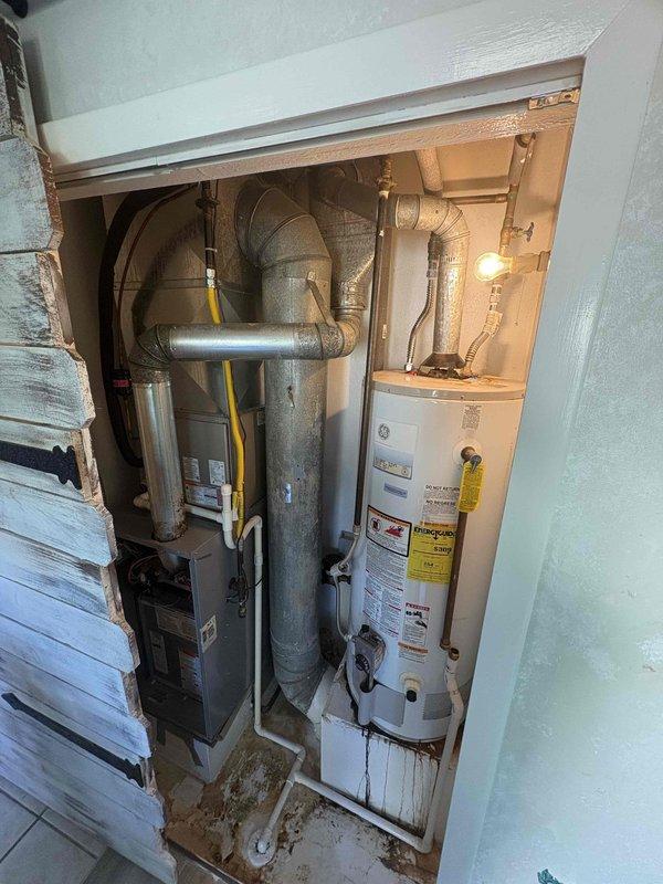Performed an on-site evaluation for HVAC system replacement of a closet-installed furnace/air handler with galvanized flue and ductwork, adjacent to a gas-fired water heater. Documented significant staining, mineral deposits, and moisture damage around the PVC condensate drain line and surrounding floor and door trim, indicating prior leakage or a condensate drainage deficiency. Replacement scope should include correcting condensate drainage and verifying flue, ductwork, and gas line connections.