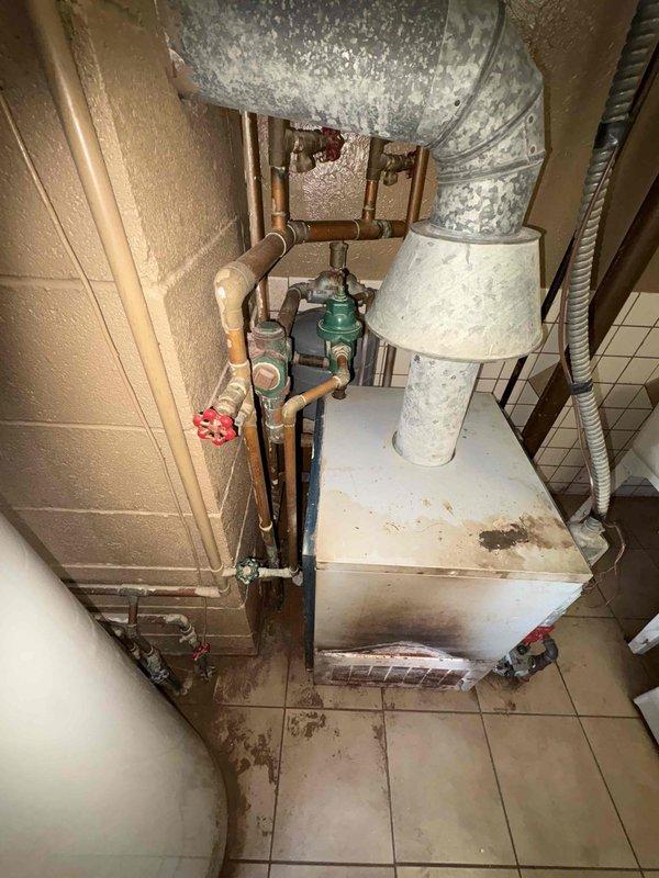Performed an on-site assessment for a boiler replacement, inspecting the existing gas-fired boiler, draft hood and vent connection, expansion tank, pressure regulator, supply and return piping, and adjacent water heater. Documented significant soot staining, scorching, and discoloration on the boiler's exterior and vent components, along with sediment and water staining on the surrounding floor, indicating combustion and moisture concerns. Photographed all equipment and conditions to support replacement scoping and recommendations.