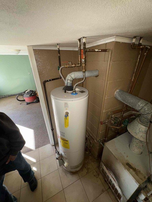 Performed an on-site assessment for a boiler replacement, inspecting the existing gas-fired boiler, draft hood and vent connection, expansion tank, pressure regulator, supply and return piping, and adjacent water heater. Documented significant soot staining, scorching, and discoloration on the boiler's exterior and vent components, along with sediment and water staining on the surrounding floor, indicating combustion and moisture concerns. Photographed all equipment and conditions to support replacement scoping and recommendations.