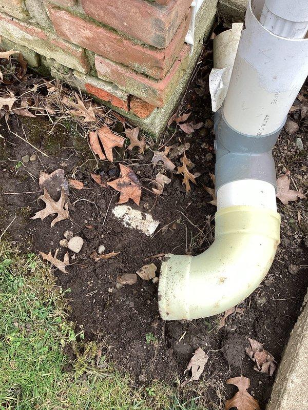 Reattached downspout to gutter system at exterior corner of brick structure. Inspected existing drainage components including PVC pipe, connector, and elbow section. Ensured proper alignment for water flow away from foundation to prevent future water damage and soil erosion.