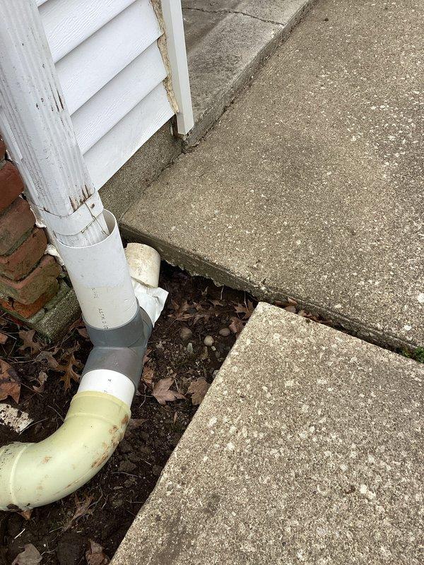 Reattached downspout to gutter system at exterior corner of brick structure. Inspected existing drainage components including PVC pipe, connector, and elbow section. Ensured proper alignment for water flow away from foundation to prevent future water damage and soil erosion.