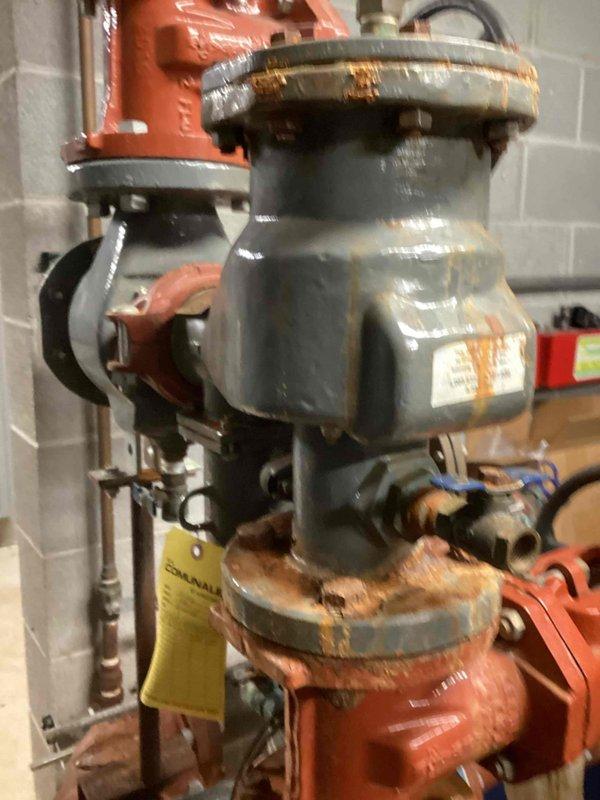 Conducted inspection of backflow prevention device located on cinderblock wall in utility room. Observed significant rust and corrosion on multiple connection points and flanges, particularly affecting the red valve wheel assembly and brass fitting components. Documented condition with photos for quote preparation on necessary repairs to bring system back into compliance.