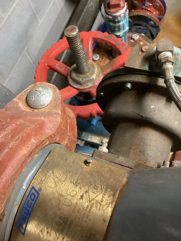 Conducted inspection of backflow prevention device located on cinderblock wall in utility room. Observed significant rust and corrosion on multiple connection points and flanges, particularly affecting the red valve wheel assembly and brass fitting components. Documented condition with photos for quote preparation on necessary repairs to bring system back into compliance.