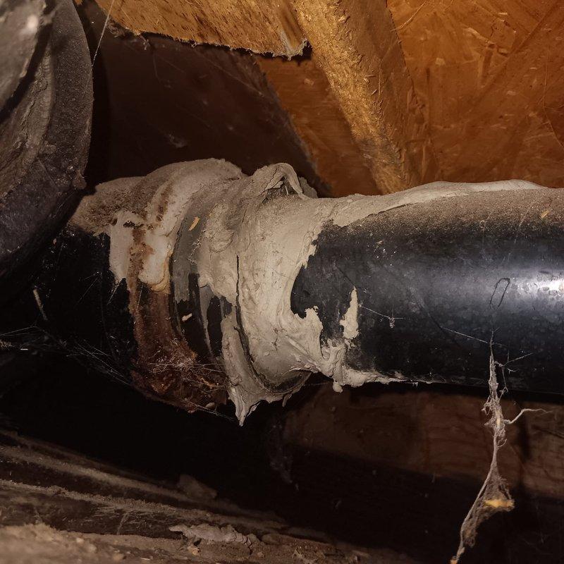 Responded to callback for active water leak from toilet penetrating through floor into kitchen ceiling below. Upon inspection, located source of leak at deteriorated toilet flange connection in second-floor bathroom. Discovered damaged wax ring seal and compromised drain pipe coupling beneath toilet, allowing wastewater to bypass plumbing system and seep through subfloor into kitchen area below. 

Accessed crawl space to assess extent of water damage and verify leak path. Found makeshift repair coupling connecting white PVC drain line to black ABS pipe with metal hose clamps, which had failed and was actively leaking. Removed temporary coupling and installed proper ABS to PVC transition coupling with approved primer and cement per code requirements. Reset toilet with new wax ring seal and closet bolts, ensuring proper flange engagement and watertight connection.

Documented deteriorated ductwork insulation in attic space near plumbing penetration, noting severe degradation of vapor bar