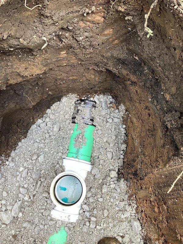 Excavated trench along right side of residential structure to access and replace damaged sewer lateral line. Removed approximately 10 feet of deteriorated piping and installed new PVC sewer pipe with proper fittings and connections. Laid pipe on crushed stone bedding material for adequate support and drainage. Installed exterior cleanout access point with green riser pipe and white cap for future maintenance accessibility. Performed camera inspection of remaining sewer line to verify system integrity and identify any additional issues downstream. Backfilled excavation upon completion of repairs.