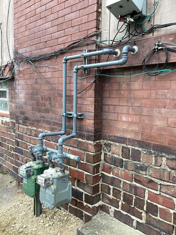 Completed gas meter relocation from exterior wall mounting to new position. Relocated two large capacity gas meters with associated supply piping, reconfigured gray piping network with new vertical and horizontal runs secured with wall-mounted brackets, and installed necessary elbows and T-joints to ensure proper gas flow to building. Modified interior basement piping connections including black steel and galvanized pipe sections with proper fittings and support clamps to accommodate the meter repositioning while maintaining code compliance and system integrity.