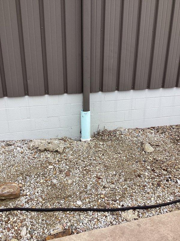 Responded to water intrusion issue at industrial property caused by cracked underground drain pipe collecting roof water. Inspected exterior building perimeter where corrugated metal wall meets cinder block foundation, identifying compromised drainage system with vertical pipe extending through gravel bed. Documented existing conditions of drain pipe infrastructure and surrounding area to assess extent of damage and determine appropriate repair strategy for the failing underground drainage system.