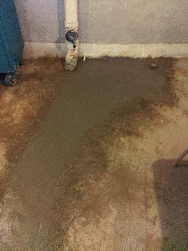 Completed comprehensive assessment of basement main drain system experiencing significant water damage and moisture infiltration. Documented extensive floor staining and discoloration patterns indicating chronic drainage issues. Photographed existing plumbing configuration including vertical drain pipe, valve connections, and areas of water accumulation to provide detailed estimate for main drain line replacement and waterproofing solutions.