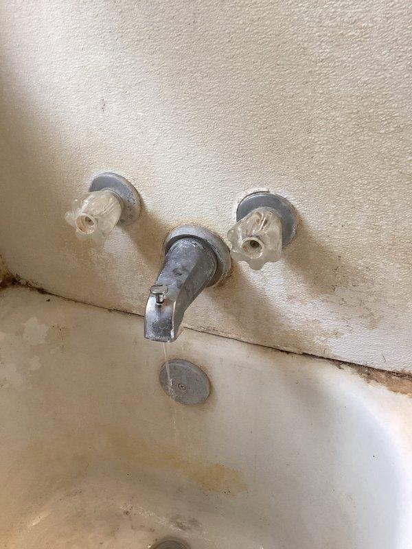 Installed customer-supplied shower valve in bathroom. The three-piece wall-mounted assembly includes a center spout and two side control valves with ceramic handles, all mounted to the tub surround wall above the bathtub fixture.