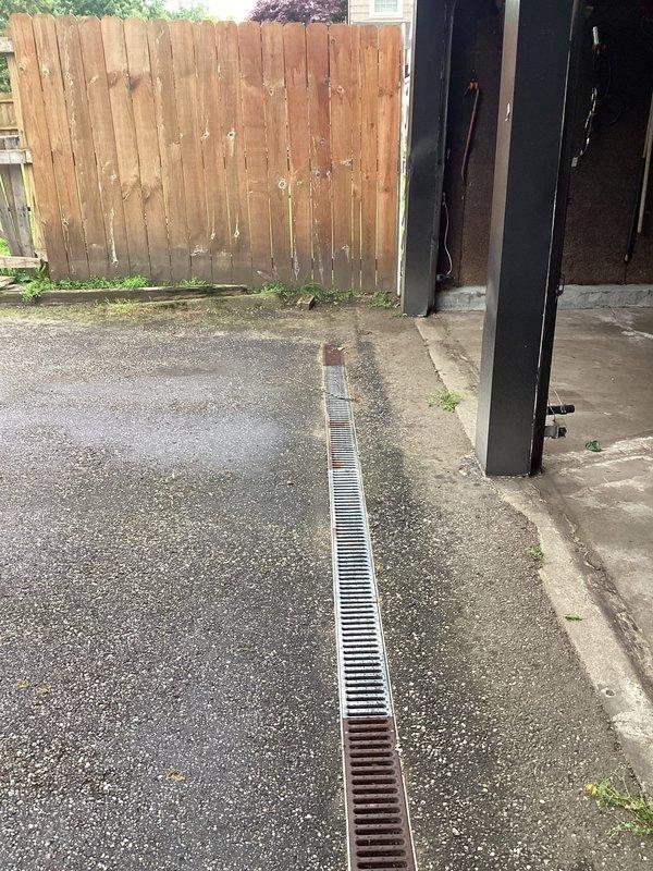 Responded to drainage issue where water from clogged garage floor drain was leaking into basement through an existing structural opening between the two spaces. Performed diagnostic assessment of the garage drainage system, identifying blockage in the linear drain grate located at the garage entrance. Documented conditions of concrete surfaces showing weathering, vegetation growth in cracks, and water intrusion pathways. Inspection revealed the garage floor drain required clearing to prevent continued water accumulation and subsequent basement infiltration through the compromised barrier between levels.