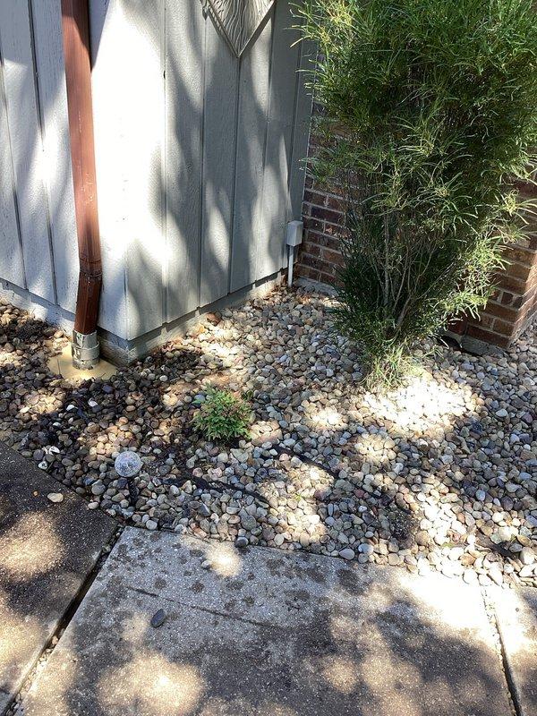 Conducted inspection of residential ground water drainage system to diagnose backflow issue causing water to enter garage during heavy rainfall events. Assessed existing garage floor drain configuration and surrounding yard grading. Documented current drainage infrastructure including exterior drainage points, downspout discharge locations, and gravel drainage areas adjacent to structure. Evaluated soil saturation patterns and potential contributing factors to reverse flow conditions in existing drain lines. Photographed site conditions showing exterior utility connections, HVAC equipment placement on concrete pad with surrounding landscape rock, foundation drainage areas, and vegetation overgrowth near building perimeter. Identified need for additional yard drainage solution to mitigate stormwater backup into garage floor drains during peak precipitation events. Prepared recommendations for installation of supplementary ground water drain system to redirect excess surface water away f