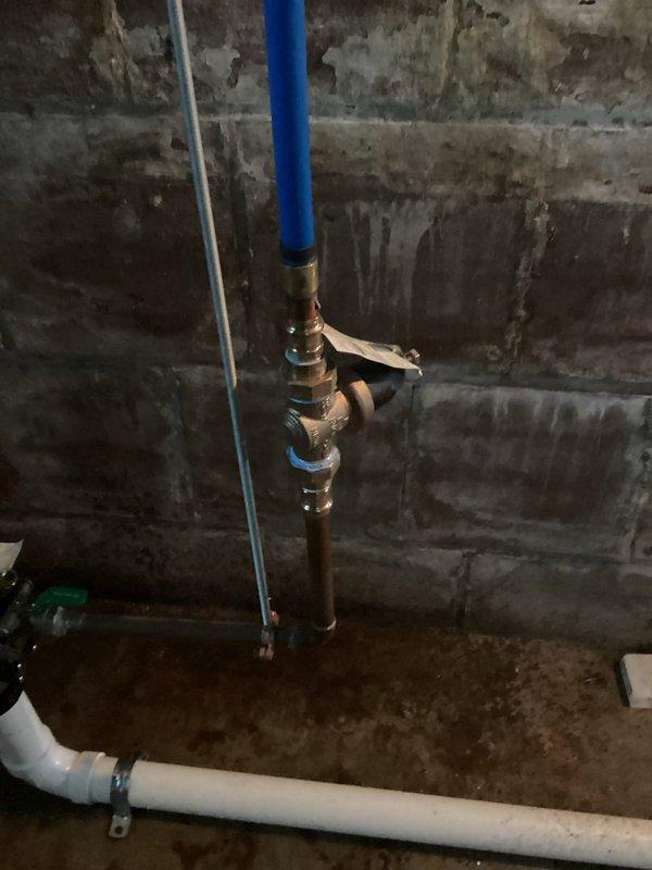 Responded to water leak in basement of 33-year-old home. Upon inspection, identified compromised backflow prevention valve assembly with brass fittings showing signs of age-related deterioration and moisture intrusion. The concrete block walls exhibited extensive water staining and discoloration, indicating chronic moisture issues. Assessed the vertical blue PVC supply line connection to the brass valve mechanism and horizontal white PVC drainage system. The copper pipe segment below the valve assembly showed corrosion consistent with prolonged water exposure. Floor surfaces displayed significant water damage and staining patterns suggesting active or recent leakage. Documented existing plumbing configuration including backflow preventer, manifold connections, and associated piping systems. Recommended valve replacement and evaluation of drainage system to prevent future water intrusion in the basement utility area.