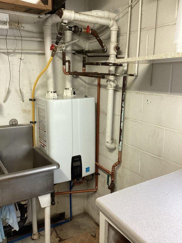 Removed existing 30-gallon conventional storage tank water heater from basement utility room and completed full installation of new Energy Star-rated tankless water heating system. Established all necessary connections including hot and cold water copper supply lines, PVC venting system routed through exterior wall with proper termination and mesh screening, flexible yellow CSST gas line supply, and electrical service to digital control panel. Installed required shut-off valves with red handles on water supply lines for code compliance and future serviceability. Configured exterior venting with dual intake/exhaust penetrations through painted concrete block wall to ensure adequate combustion air supply and proper exhaust discharge. All plumbing connections tested and verified leak-free, unit powered up and programmed to optimal temperature settings, system fully operational upon completion.