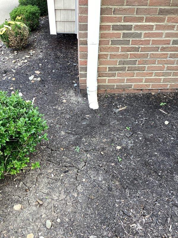 Completed assessment of residential downspout system and drainage conditions at brick home exterior. Inspected white-painted downspout running along foundation wall, evaluated water discharge patterns, and examined surrounding landscaping and mulch beds for signs of water damage or improper drainage. Documented current condition of downspout installation, foundation perimeter, and ground surface materials to provide comprehensive quote for necessary repairs or improvements to gutter drainage system.