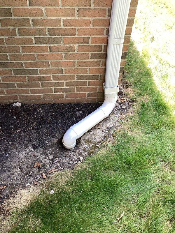 Conducted assessment of exterior downspout and drainage system at brick building foundation. Inspected white PVC downspout assembly including vertical runs, elbow joints, and horizontal extension components for proper water diversion away from foundation. Documented current condition of drainage infrastructure and landscaping interface where downspout terminates in mulched bed area. Photographed installation for quote preparation regarding necessary repairs to downspout system.