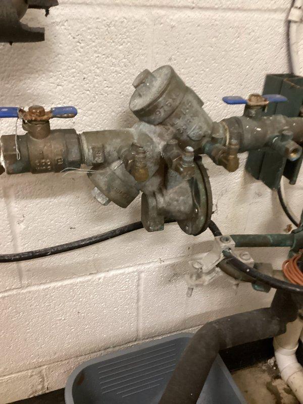 Responded to water leak at main water line valve assembly located near wash sink area. Upon inspection, identified steady drip originating from aged plumbing assembly mounted on utility room wall, consisting of ball valves with blue handles, pressure regulator, water meter, and multiple threaded pipe connections showing significant weathering and oxidation. Documented existing conditions with detailed photographs of the complete valve assembly, backflow preventer, and associated piping components for assessment. Assembly displayed greenish-gray patina on brass and copper fittings, indicating long-term exposure to moisture. Prepared two required quotes as specified: first for like-for-like repairs to address the leak at the main valve connection, and second for comprehensive system evaluation. Photographs captured all plumbing components including valve positions, meter assembly, electrical junction box connections, and drain pan placement for proposal approval and documentation purpose