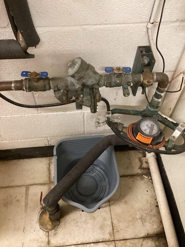 Responded to water leak at main water line valve assembly located near wash sink area. Upon inspection, identified steady drip originating from aged plumbing assembly mounted on utility room wall, consisting of ball valves with blue handles, pressure regulator, water meter, and multiple threaded pipe connections showing significant weathering and oxidation. Documented existing conditions with detailed photographs of the complete valve assembly, backflow preventer, and associated piping components for assessment. Assembly displayed greenish-gray patina on brass and copper fittings, indicating long-term exposure to moisture. Prepared two required quotes as specified: first for like-for-like repairs to address the leak at the main valve connection, and second for comprehensive system evaluation. Photographs captured all plumbing components including valve positions, meter assembly, electrical junction box connections, and drain pan placement for proposal approval and documentation purpose