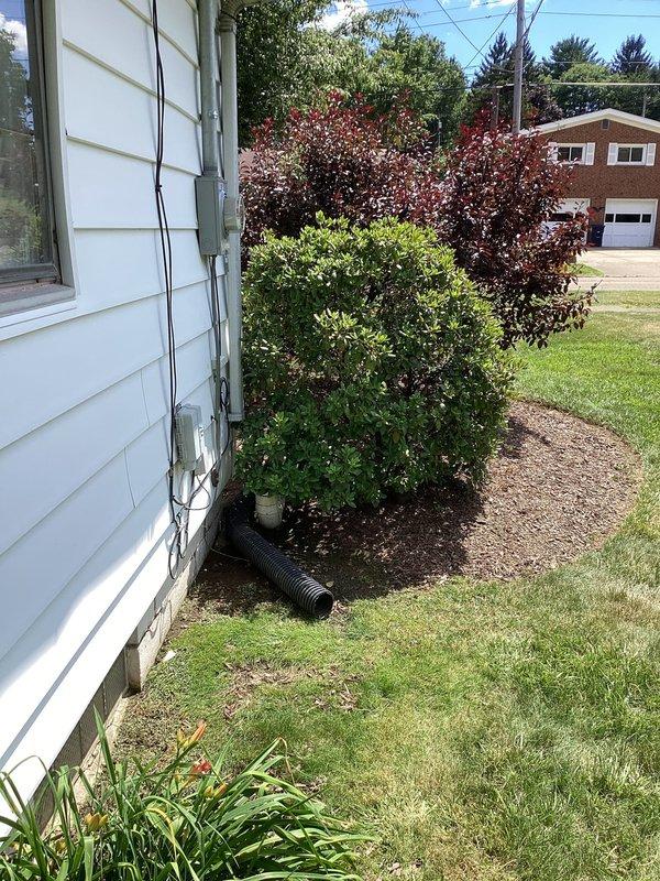 Responded to service call for improperly functioning downspout drainage system at hillside residential property. Upon inspection, found existing downspout extension had been previously compromised and temporarily repaired, resulting in inadequate water diversion. Due to property's slope toward the foundation, water was draining toward the house instead of away from the structure, creating potential foundation damage risk.

Corrected downspout routing to redirect water flow away from building foundation toward the roadway. Installed proper drainage extension with black corrugated pipe to ensure adequate water conveyance down the slope. Verified proper grade and positioning to prevent water accumulation near foundation plantings and building perimeter. System now functions as designed to protect structural integrity during precipitation events.