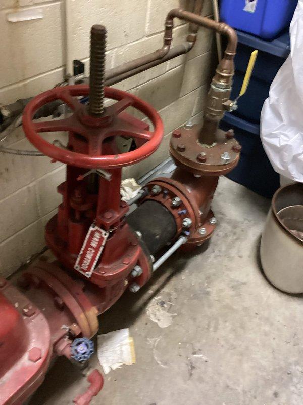 Conducted on-site assessment of existing plumbing infrastructure to provide estimate for backflow preventer installation. Inspected main water supply system including large gate valve assembly with red handwheel, connecting copper distribution piping, and associated mechanical components mounted against cinder block wall in mechanical room. Documented current configuration of water supply lines, valve conditions showing age-related wear and corrosion, and existing backflow prevention equipment. Photographed valve assembly, copper piping network, pressure regulation components, and floor drain access points to support accurate estimation of backflow device installation requirements and necessary modifications to existing plumbing system.