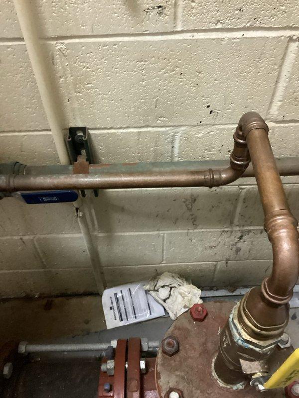 Conducted on-site assessment of existing plumbing infrastructure to provide estimate for backflow preventer installation. Inspected main water supply system including large gate valve assembly with red handwheel, connecting copper distribution piping, and associated mechanical components mounted against cinder block wall in mechanical room. Documented current configuration of water supply lines, valve conditions showing age-related wear and corrosion, and existing backflow prevention equipment. Photographed valve assembly, copper piping network, pressure regulation components, and floor drain access points to support accurate estimation of backflow device installation requirements and necessary modifications to existing plumbing system.