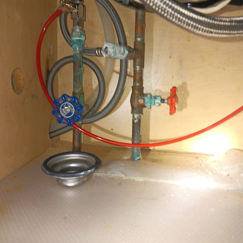 Responded to commercial water leak and low water pressure complaint. Upon inspection, identified corroded fittings on copper piping at water heater location. Examined water softener system and associated plumbing connections. Found turquoise-colored pipe fitting showing significant corrosion and mineral deposit buildup, contributing to reduced water flow. Inspected shut-off valves, braided stainless steel supply lines, and drain pan beneath water heater assembly. Confirmed presence of deteriorated fittings requiring replacement to restore proper water pressure and prevent potential leaks. Documented plumbing infrastructure condition including blue and red-handled valves, flexible metal hoses, and mounting configuration within utility enclosure.