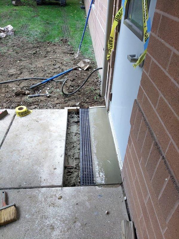 Installed trench drain system at building exterior doorway entrance per specifications. The drainage channel with black perforated grate was set into freshly poured concrete at the threshold to manage water runoff and prevent pooling at the entryway. Work area was secured with caution tape during concrete curing process.